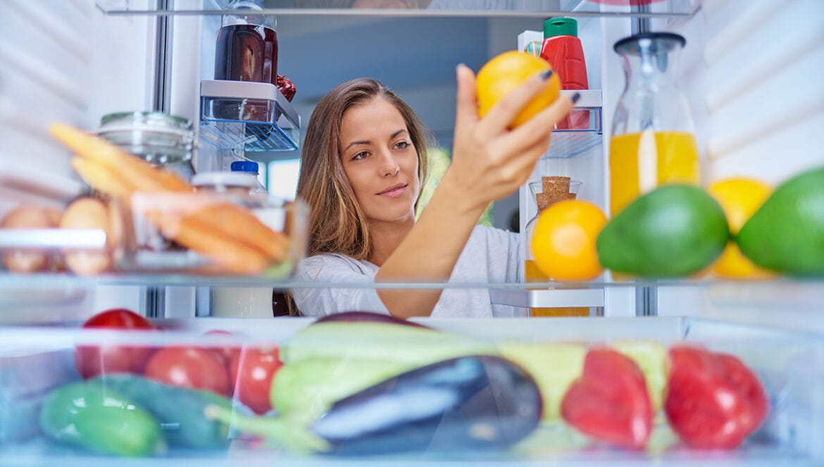 Mujer mira interior de refrigerador y revisa la temperatura de una naranja sosteniéndola con la mano
