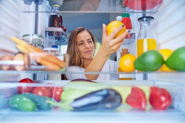 Mujer mira interior de refrigerador y revisa la temperatura de una naranja sosteniéndola con la mano